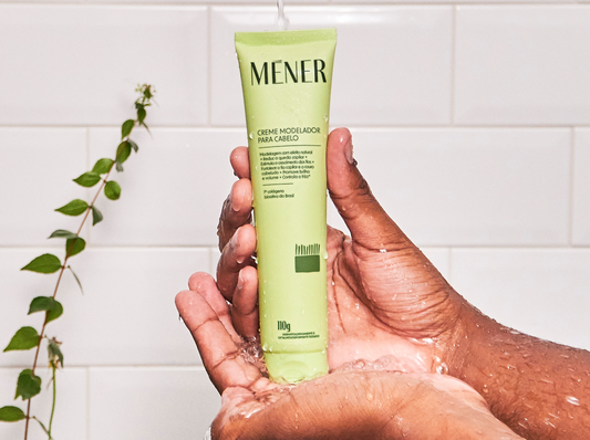Mão segurando o creme modelador para cabelo Méner no banho, com água escorrendo.